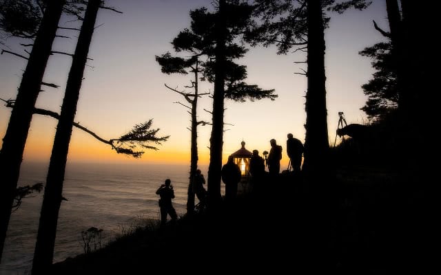 ON1 photographers with tripods capturing a coastal lighthouse silhouetted against an orange sunset