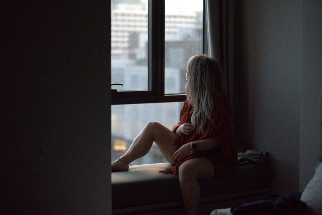 Before: Woman sitting by a window in a dim room looking out at the city