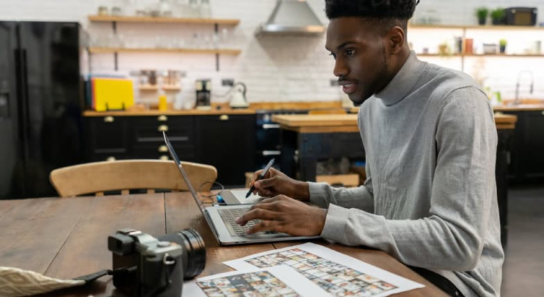 Photographer reviewing photo contact sheets while editing images on a laptop at a kitchen table, with a DSLR camera and printed proofs nearby