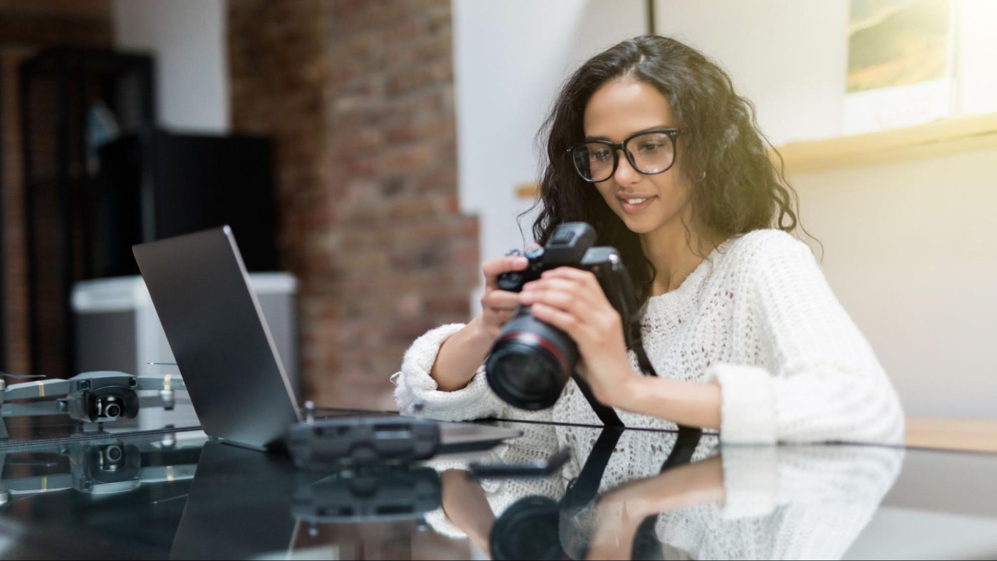 Woman reviewing photos on a DSLR camera while sitting at a desk with a laptop and drone equipment.