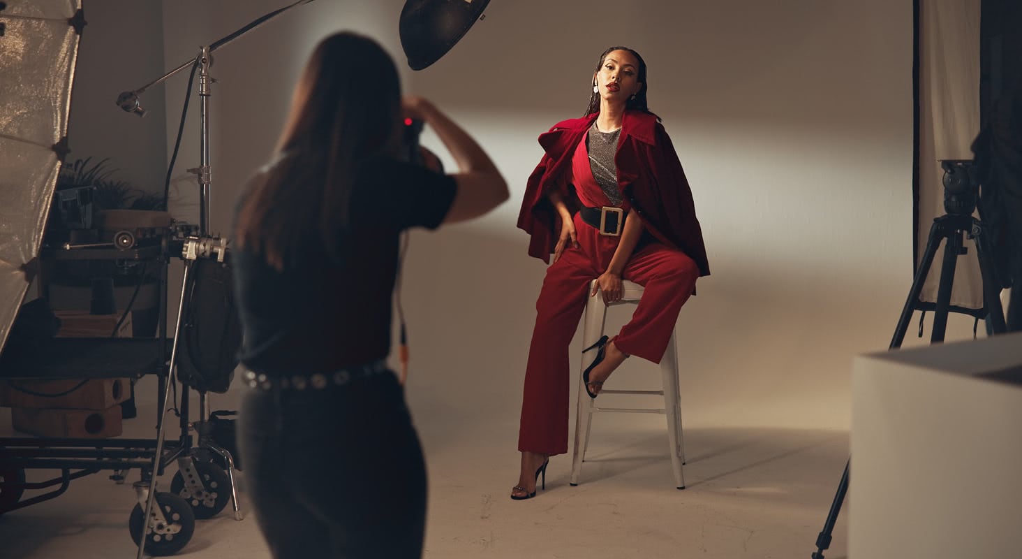 Behind-the-scenes fashion studio shoot with a model in a red outfit posed on a stool under studio lighting