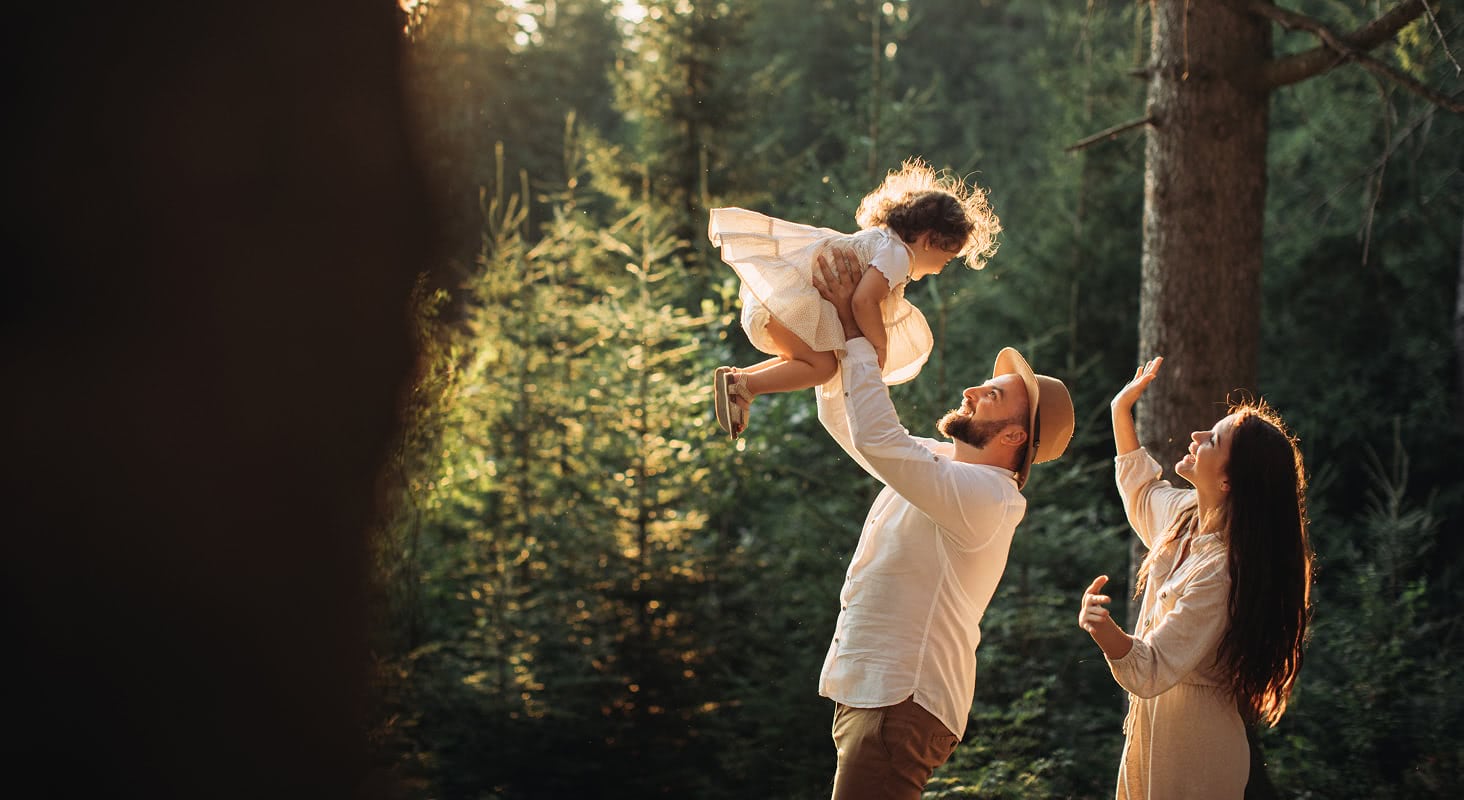 Father lifting a young child into golden sunlight in a forest while mother watches and smiles