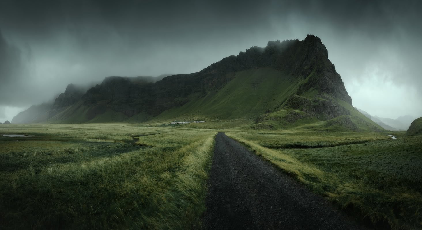 Moody dirt road leading toward a dramatic green mountain peak under dark overcast skies