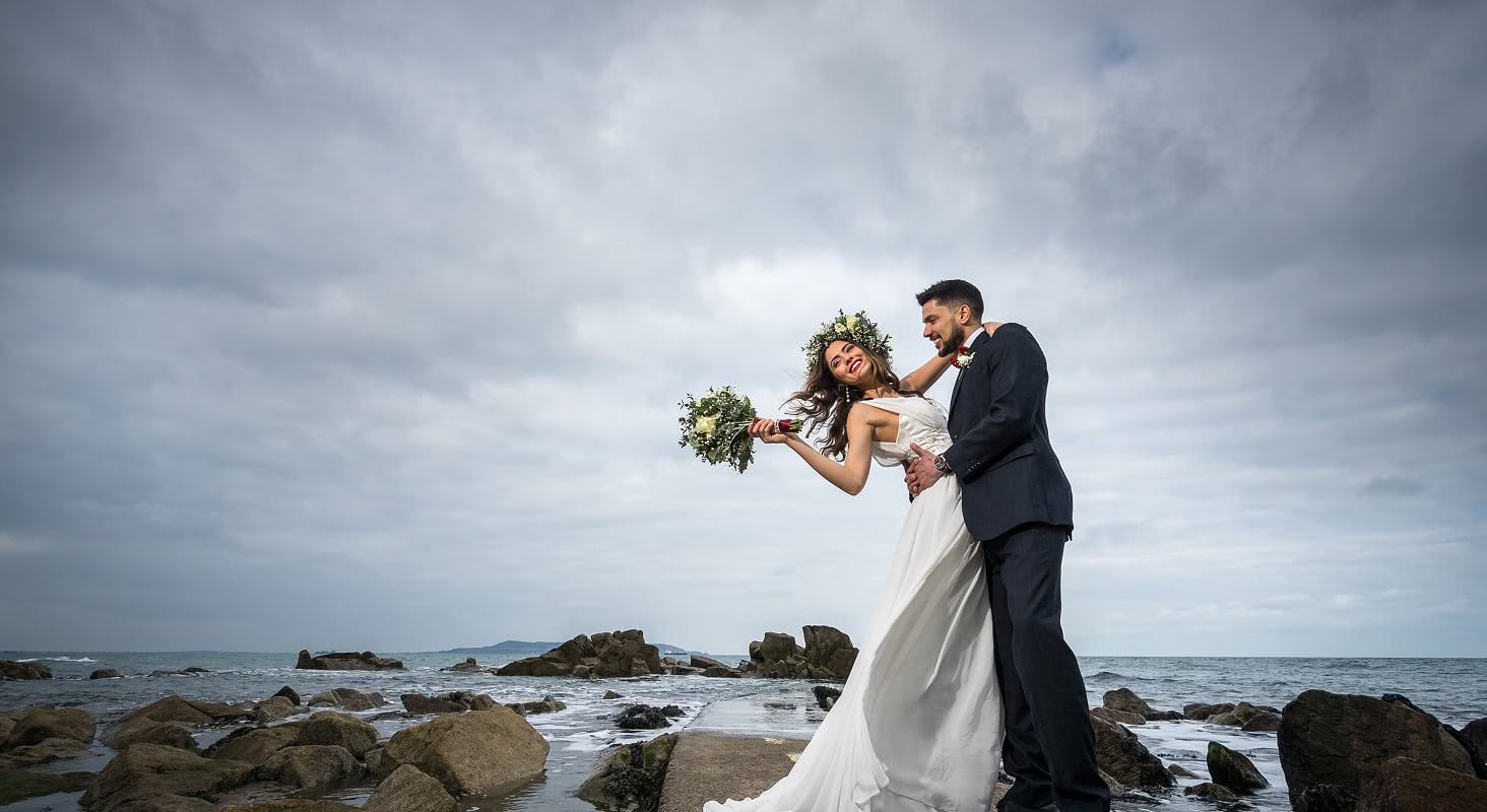 Bride and groom embracing on a rocky ocean shoreline with a cloudy sky and bouquet in hand