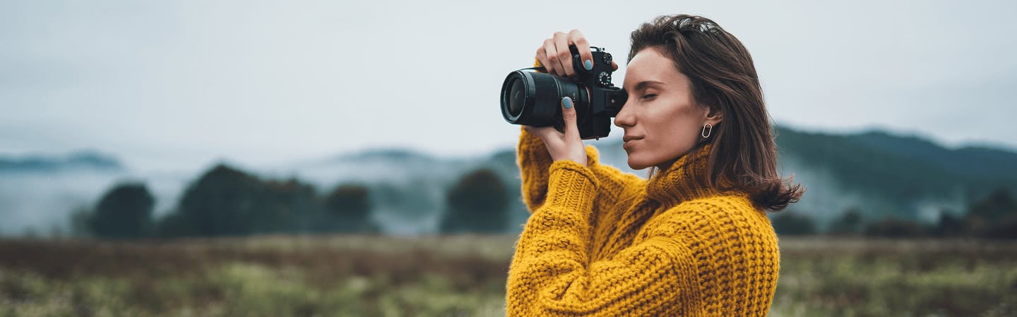 Photographer in a yellow sweater shooting with a mirrorless camera against a misty mountain landscape.