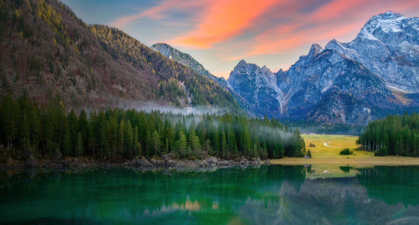 A misty alpine lake below snowy peaks under a pink sunset sky